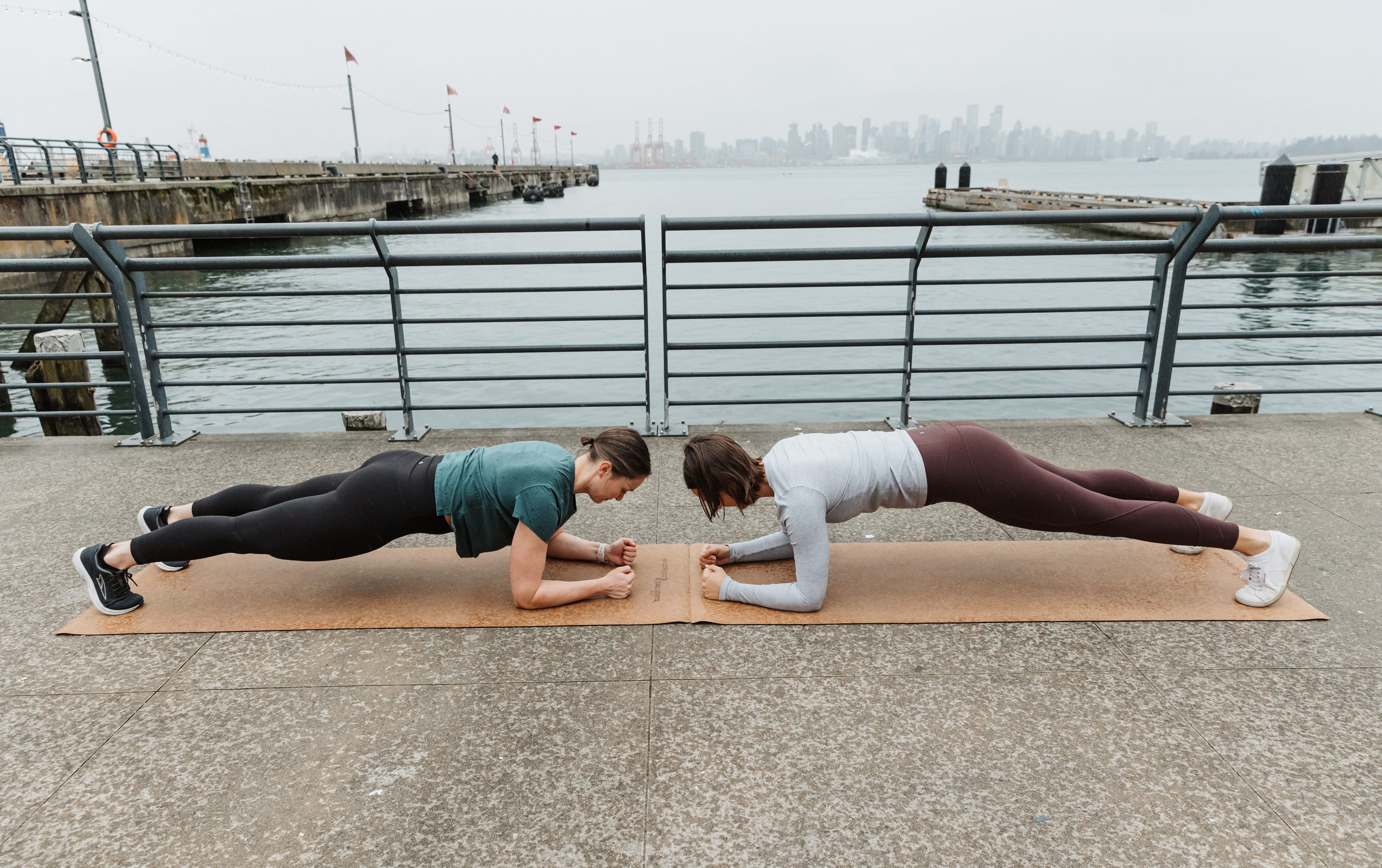 Briana and Jess facing each other in plank position. They are on a dock by the water with the Vancouver skyline in the background.