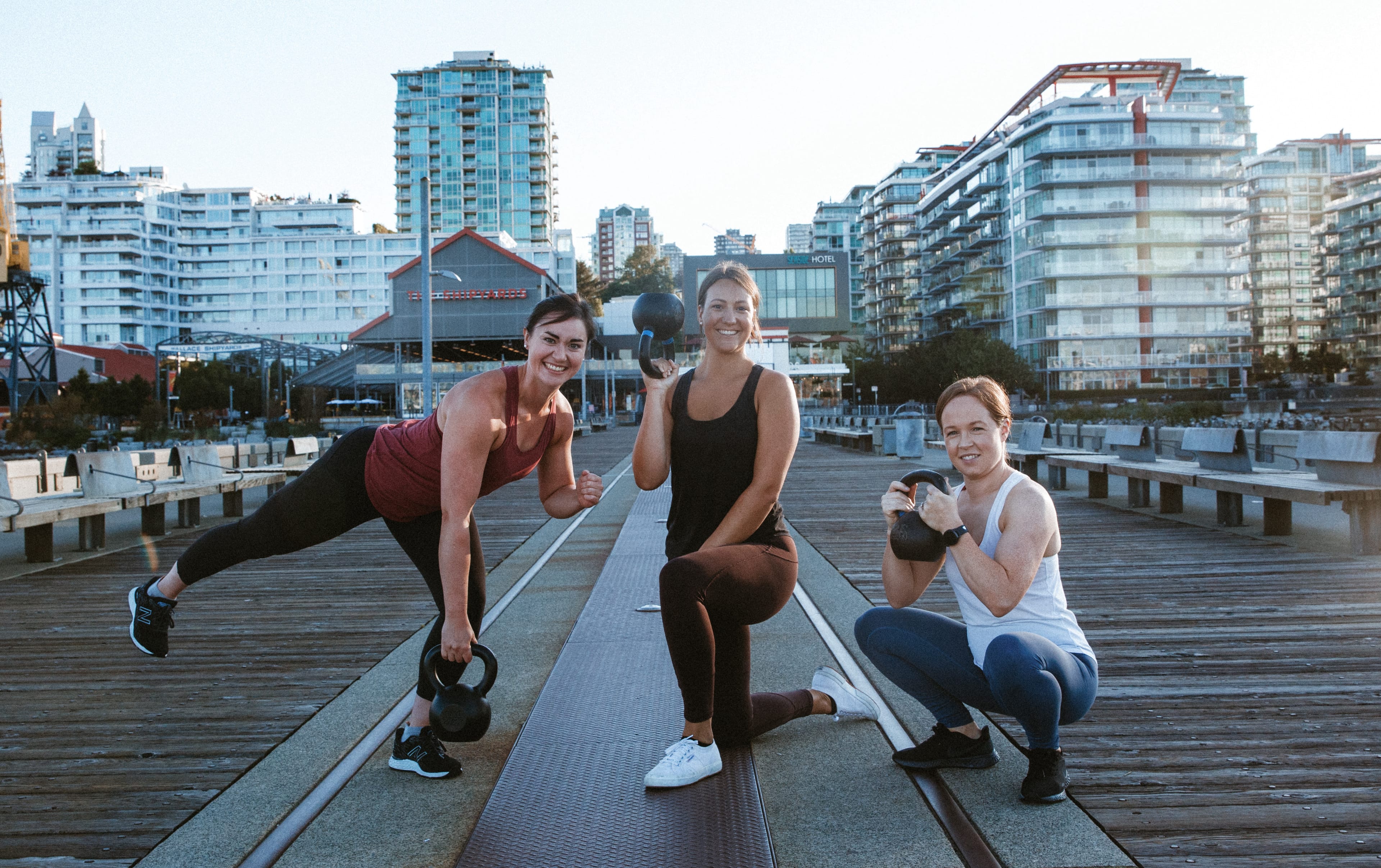 Andrea, Briana, and Jess each doing kettlebell exercises on a dock by the water, with the North Vancouver skyline in the background.